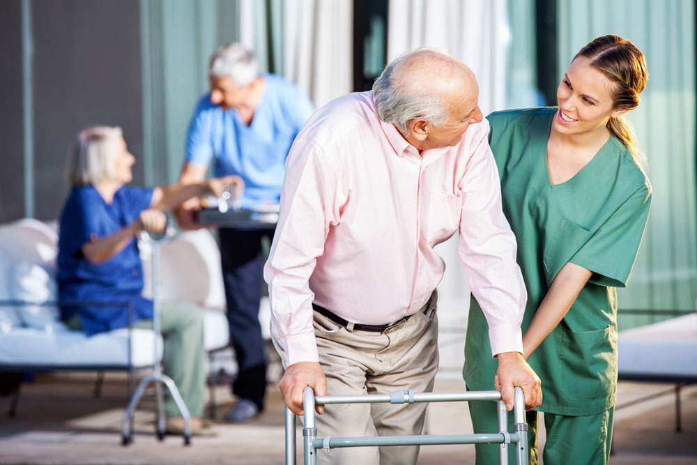 Happy female caretaker assisting senior man in using Zimmer frame at nursing home yard